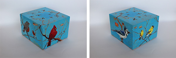 A cube model with a baby blue mottled background. Line drawn trees of shades of rust red and brown are the perch for different birds on each side: a pair of yellow finch, a bluejay, a bold red cardinal and a pair of robin with red breasts.