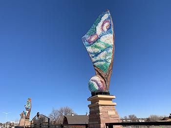 Blue clear sky with an abstract sculpture at center. Sculpture is fin shaped with white, purple, blue and green colors splashed across it.