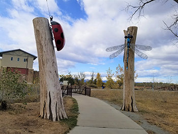 Concrete bike path with two large insect sculptures flanking it, each elevated on a tall wooden post. A ladybug to the left and a blue lacy winged dragonfly to the right.