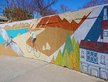 Painted wall depicting a stylized Longmont. A bicyclist rides on a path past a lake, mountains and a hawk to his left with buildings and pine trees behind him.