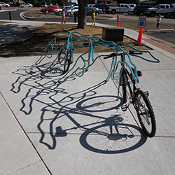 a photo of a bike rack that are made from simple lines resembling swimmers is shown with a few bikes attached. The shadows of the rack is shown on the bottom where the sunlight hits the pavement.