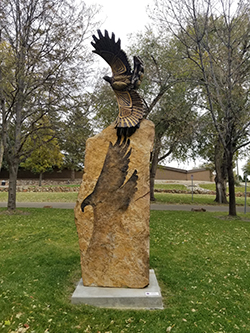 a sculpture of a bronze hawk on a large stone pedestal is pictured in a park with green grass and trees. the stone base has a superimposed shadow of the bird and its wingspan. the bird itself has geometric designs carved into its body.