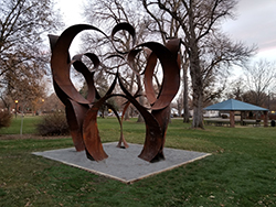 a photo of an abstract steel sculpture with many loops and circles connected to create a gazebo like structure. a parkcan be seen in the background with trees and the sculpture is sitting on a concrete pad. a shelter with a green roof is in the distance and grey overcast sky with clouds.