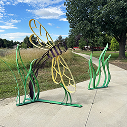 a photo of a bike rack that is also shaped like grass blades and a black and yellow dragonfly on a sidewalk with trees and a blue sky with clouds in the background.