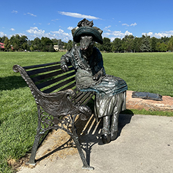 a view of a giraffe-human woman bronze sculpture sitting on a bench with trees and a blue sky behind it. The giraffe-person wears clothing typical of a 1900's woman with frilly skirts and a wide brimmed hat.