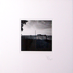 a black and white photograph with a white mat border is pictured of a landscape featuring a gate and a road and a cloudy sky in the distance with some tree leaves on the left corner of the image. the mat is signed by the artist in pencil at the bottom right of the photo.