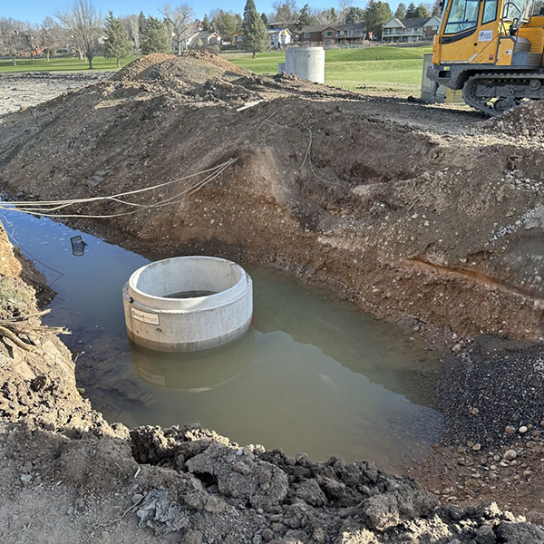 A hole in the ground is partially filled with water and large irrigation pipe parts.