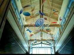 a view of a mobile installation is shown from the ground looking up to the ceiling at the Longmont Recreation center. The mobile consists of colorful swiling shapes as well as black and white striped shapes. 