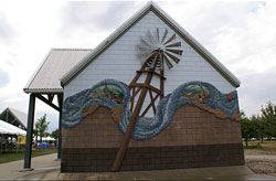 the side of a small building is shown with a textured façade of a mosaic water wave with a tall windmill in the center. The sky above has many clouds and the background has some trees and other buildings far away. 