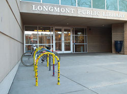 The entrance to the Longmont Library is pictured with doors in the center of the image. colorful painted u shaped bike racks are on the left side of the image. 