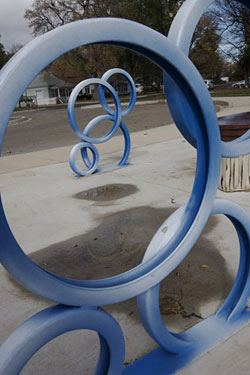 an up close image of a bike rack with another similar bike rack is shown inside. The bike rack is blue and looks like round bubbles. The concrete sidewalk is also shown with trees in the background.. 