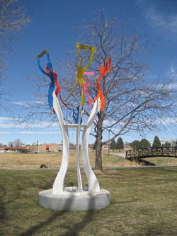 an image of a metal sculpture is shown with colorful simplified figures branching out at the top. A park is seen in the background with a brown bridge, leafless trees, and a blue sky.