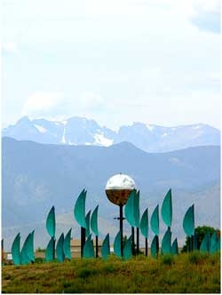 an image of a large sculptural installation outside is shown with a central high shine orb with green leaf like protrusions around the orb. The Colorado front range mountains as well as a cloudy sky are seen behind. 