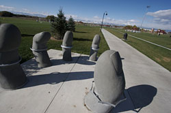 an image of a bike rack that appears as fingers emerging from the concrete sidewalk. The perscpective of the photo shows a sidewalk disappearing into the distance along the right side of the sculpture with blue sky and clouds in the background. 