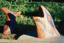 a sculpture that resembles a fish with red clay and mosaic tiling is emerging from a concrete sidewalk. Grass is seen behind the fish sculpture.