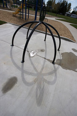 an abstract bike rack is shown with multiple legs. the concrete ground beneath has some rain puddles and a children's playground is behind the sculpture. 