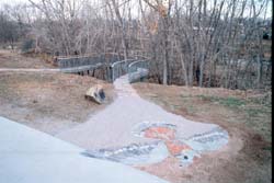 an outdoor art installation featuring a mosaic bird and a swooping concrete walkway amongst trees and green grass