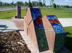 an angular stone and mosaic sculpture is pictured in a park with a sidewalk running through the sculpture. Trees, a street and grass are in the background. 