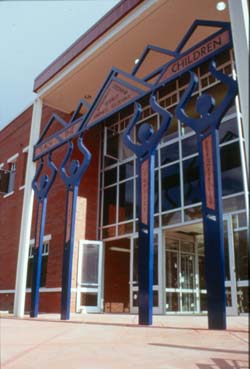 the boulder county courthouse entrance is shown with a towering metal sculpture of simplified figures holding up triangular mountains shapes and text that read Teach the Children.