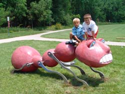 A photo of two young children are seen on top of a comically large red ant with teeth. Behind them is a sidewalk, trees, and grass.