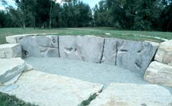 a recessed stone area with carvings of plant life are shown with a gravel pit and green grass with a treeline and a blue sky