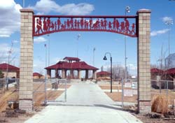 an entrance to a park is shown with brick columns and a red metal arch with silhouettes of baseball figures is shown. Behind, a shelter with a red roof can be seen. Blue skies with clouds is above. 