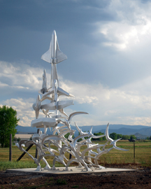 a silver airplane sculpture comprised of many small airplane objects are pictured with many at the base leading from many to just one airplane figure facing the sky. The airport is seen behind the sculpture as well as a blue sky with clouds and some mountains.