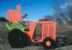 a cartoon cut out sculpture of a bright red turkey wearing a bonnet is riding a red tractor with a green seat and black wheels. The sculpture is shown in a park field with a blue sky and some trees in the background.