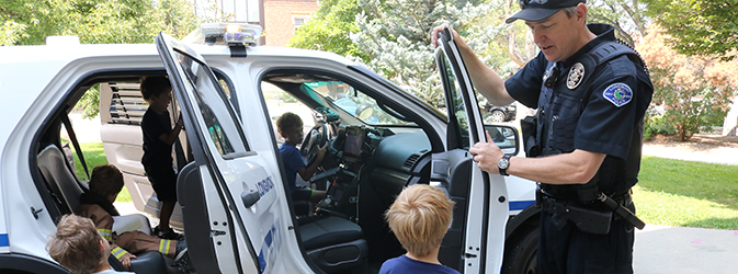 A male police officer shows elementary school children a police car while they climb inside