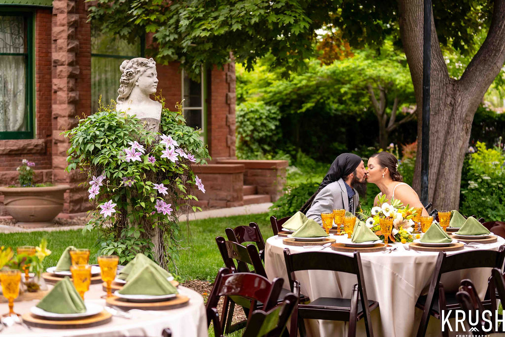 Couple share a kiss at a party table in front of the Callahan House. Statues and blooms are in the garden around them.