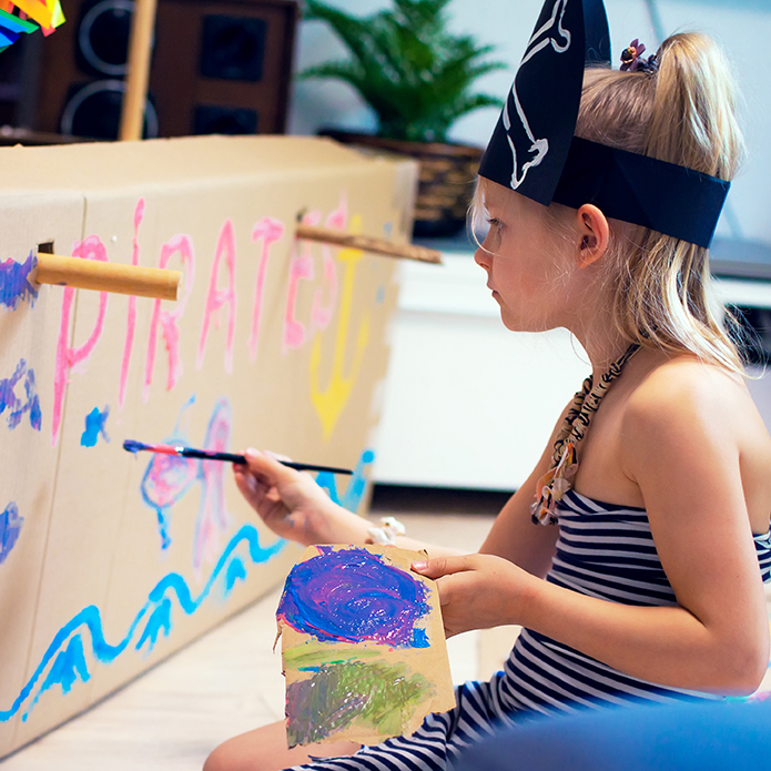 A girl in a pirate costume paints a large home-made sea ship made of cardboard. The game of pirates with the whole family, creative development, dreams of adventure and travel