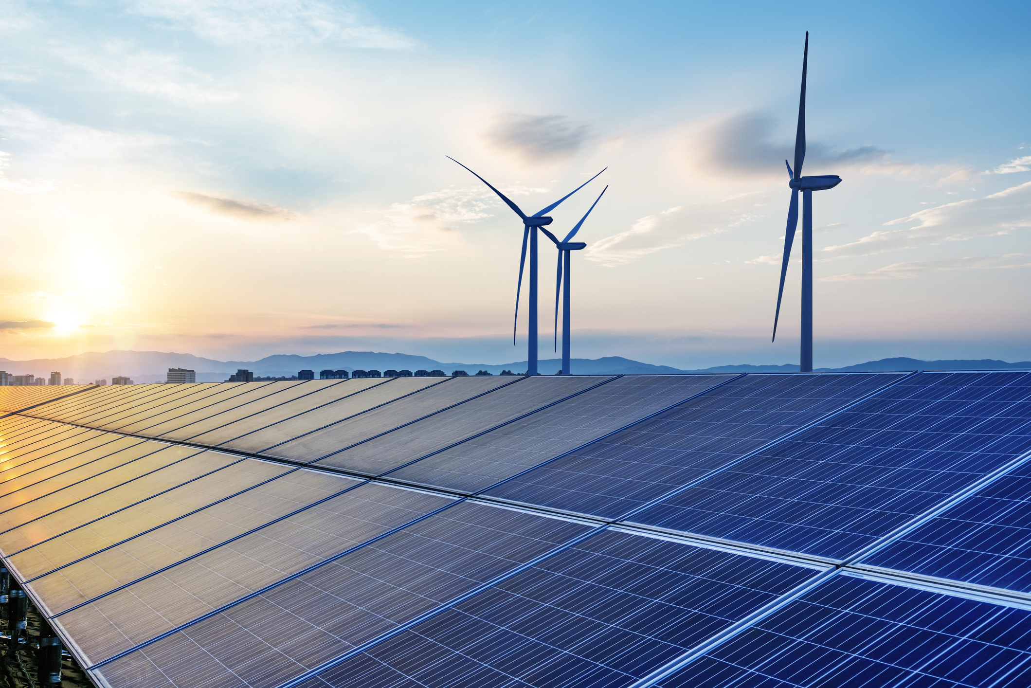 Wind turbines stand behind an array of solar panels in the sunlight