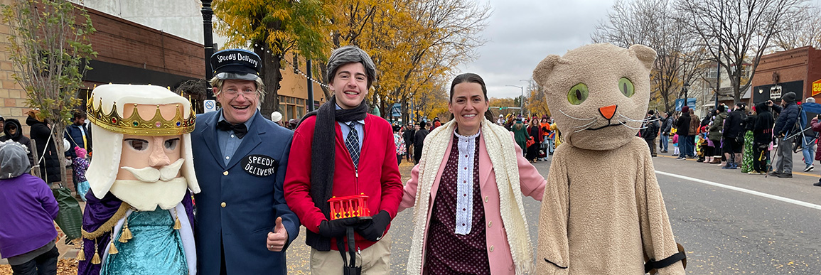 Four adults in costume with the Halloween parade and crowd watching in the background.