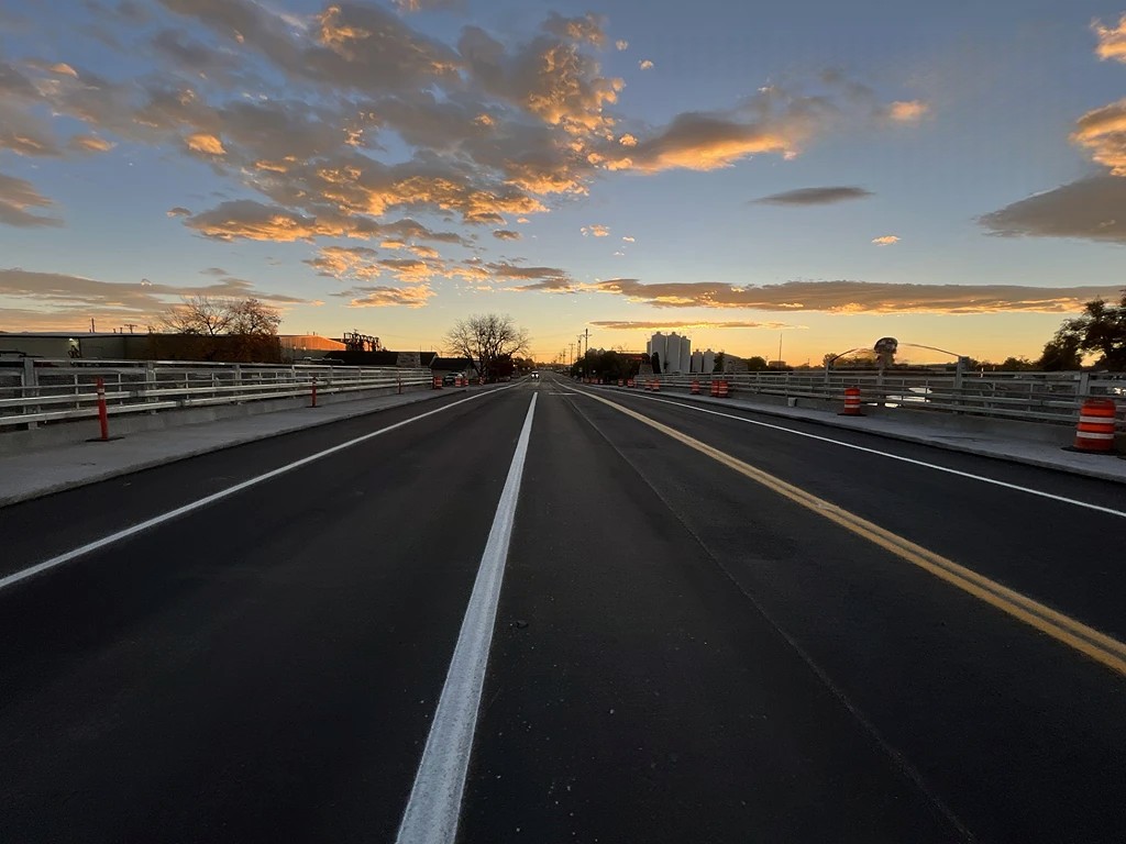 A newly paved road stretches across the Boston Ave bridge at sunset, with a clear sky above displaying scattered clouds illuminated by the warm glow of the setting sun. Orange construction barrels line the sides of the road, and industrial buildings and trees are visible in the background, silhouetted against the sky.