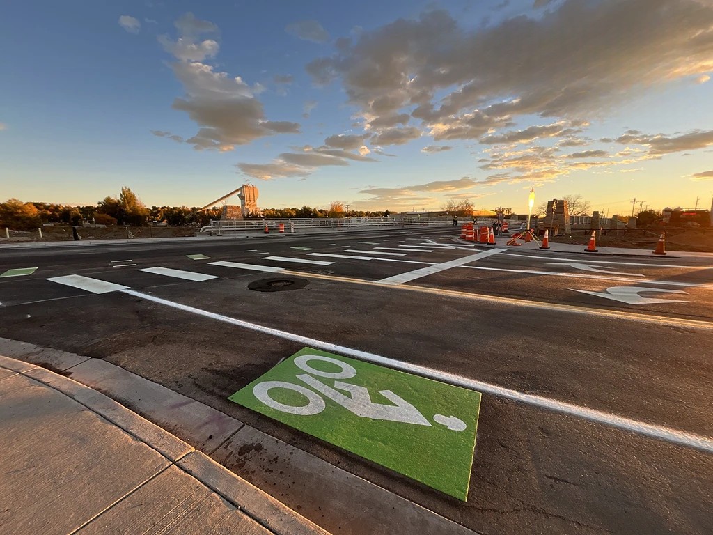 An intersection on a newly paved road at sunset, with clear lane markings, crosswalks, and green bike lane symbols. Orange construction cones and barrels are set up across the road. The sky is partially cloudy with a warm, golden glow from the setting sun, and trees and structures are visible in the background.