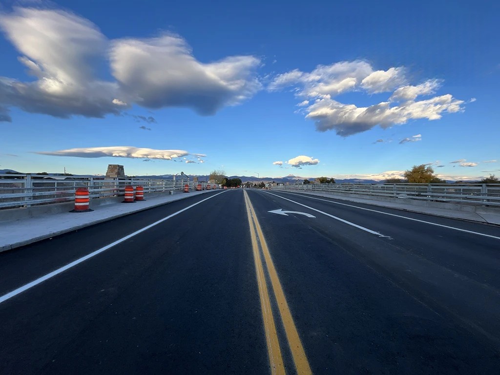 A wide, newly paved road on the Boston Ave bridge under a clear blue sky with scattered clouds. Orange construction barrels line the sides of the bridge. Mountains and trees can be seen in the background, along with a directional arrow on the road pointing left.